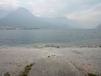 an empty bench on the beach of a lake in the background is mountains and a city in the distance