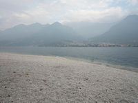 an empty bench on the beach of a lake in the background is mountains and a city in the distance