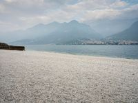 an empty bench on the beach of a lake in the background is mountains and a city in the distance