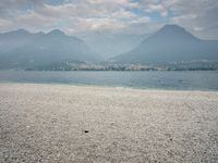 an empty bench on the beach of a lake in the background is mountains and a city in the distance