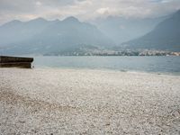 an empty bench on the beach of a lake in the background is mountains and a city in the distance