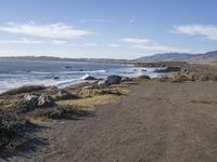 a paved dirt road with a bench and some mountains and waves in the water behind it