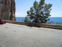 a red car parked near a cliff next to the ocean on a road side with blue skies