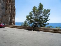 a red car parked near a cliff next to the ocean on a road side with blue skies