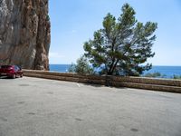 a red car parked near a cliff next to the ocean on a road side with blue skies