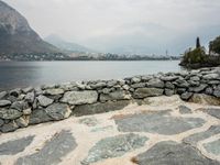 the stone wall and water are beside the mountains and buildings, which are visible in the background