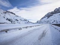 a narrow dirt road surrounded by snow covered mountains under a blue sky and clouds are visible