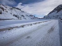 a narrow dirt road surrounded by snow covered mountains under a blue sky and clouds are visible