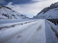 a narrow dirt road surrounded by snow covered mountains under a blue sky and clouds are visible