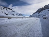 a narrow dirt road surrounded by snow covered mountains under a blue sky and clouds are visible