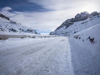 a narrow dirt road surrounded by snow covered mountains under a blue sky and clouds are visible