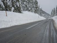 an empty road next to a snowy mountain range with fir trees along both sides of the road