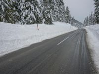 an empty road next to a snowy mountain range with fir trees along both sides of the road