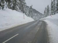 an empty road next to a snowy mountain range with fir trees along both sides of the road