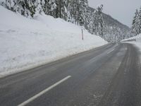 an empty road next to a snowy mountain range with fir trees along both sides of the road
