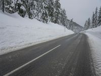 an empty road next to a snowy mountain range with fir trees along both sides of the road