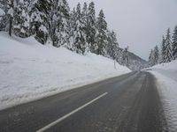 an empty road next to a snowy mountain range with fir trees along both sides of the road
