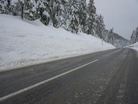 an empty road next to a snowy mountain range with fir trees along both sides of the road