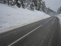 an empty road next to a snowy mountain range with fir trees along both sides of the road