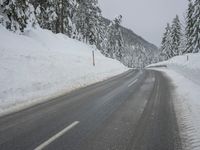 an empty road next to a snowy mountain range with fir trees along both sides of the road