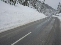 an empty road next to a snowy mountain range with fir trees along both sides of the road