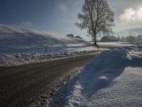 a snow covered road passing by a tree near a hill on a sunny day with a sky