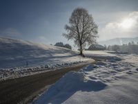 a snow covered road passing by a tree near a hill on a sunny day with a sky