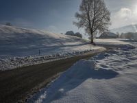 a snow covered road passing by a tree near a hill on a sunny day with a sky