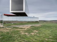 an empty glass building sits on top of grass near a field of dead grass and hills