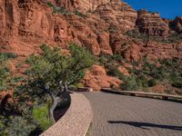 the sidewalk and red rock wall are on the side of a street near a bench with trees and bushes