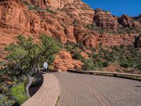 the sidewalk and red rock wall are on the side of a street near a bench with trees and bushes