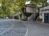 a building sitting between trees and some stairs in a street with cobblestones on it