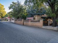 a lone street that is lined with trees, houses, and benches next to a fence