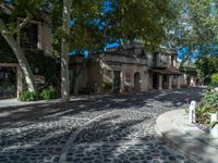 a large house sitting on a cobblestone driveway next to trees and buildings on a sunny day