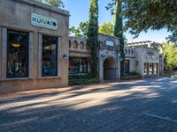 an empty city street that is surrounded by trees and plants, with two shops and one building