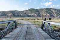 a man standing on the end of a wooden bridge near mountains and a road with no traffic or other people in it