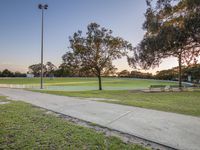 a park with an empty baseball field, tree, bench, and tennis court in the evening