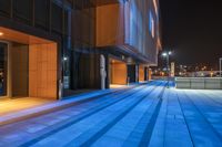 a walkway next to a very tall building at night with bright lights reflecting in the windows