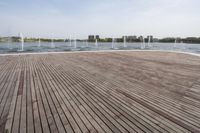 a wooden deck next to a lake surrounded by buildings and a park area with fountains