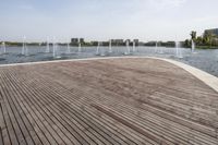 a wooden deck next to a lake surrounded by buildings and a park area with fountains