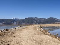 an empty dirt road is by the water with mountains in the background in this photo