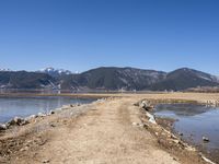 an empty dirt road is by the water with mountains in the background in this photo