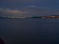 a boat sailing in the water at night with a city lights in the background on a cloudy day