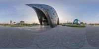 a curved sculpture sits in an empty plaza of an area of a city and looks like it's made from glass