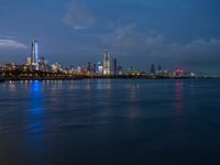 a view of the city skyline at night from across the water with clouds above it