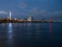 a view of the city skyline at night from across the water with clouds above it