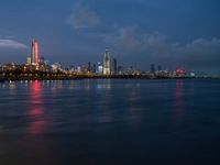 a view of the city skyline at night from across the water with clouds above it