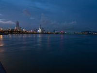 the city and skyline are at night from the water's edge in the foreground