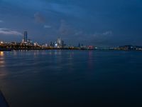 the city and skyline are at night from the water's edge in the foreground