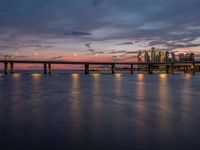 a bridge spanning over the ocean next to buildings at sunset on a pier with a moonlit sky and light reflection